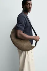 Bolso de hombro con diseño de media luna, confeccionado en gamuza color marrón. Cuenta con un compartimento principal con cierre de cremallera y un bolsillo interior con cierre. Posee un asa de hombro integrada.