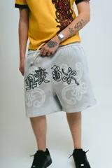Bermuda de felpa color gris claro con efecto lavado (washed). Presenta un corte ancho con cintura elástica y cordón beige. El diseño frontal incluye un estampado serigrafiado en negro con texto y motivos florales/ornamentales en bordado blanco. Posee remaches metálicos cromados en los laterales y bolsillos.