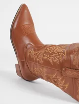 Botas texanas de caña media en color marrón, confeccionadas en cuero vegano. Presentan bordados decorativos en contraste a lo largo de la caña y en la puntera, con un taco cuadrado de madera y punta fina.
