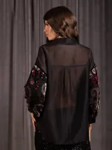 Camisa de manga larga con diseño de terciopelo estampado con motivos florales y paisley, combinada con paneles de tela transparente en la parte superior. Presenta cuello clásico, cierre frontal con botones y mangas abullonadas.