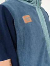 Campera de polar con capucha, cierre frontal completo y diseño en bloques de color azul y azul marino. Presenta detalles en nylon en la zona del cierre y la capucha, con logo de la marca aplicado en el pecho.