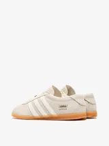 Championes Adidas Gazelle Lo Pro W de perfil bajo, confeccionados en gamuza color gris claro, con las tres tiras características en tono crudo y suela de goma translúcida. Incluye el nombre del modelo y la marca en letras doradas en el lateral.