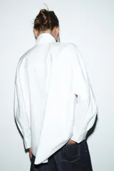 Camisa blanca de popelín con cuello subido y lazo con terminación de puntilla. Tiene manga larga con hombro caído y puños abotonados. El cierre es frontal con botones.