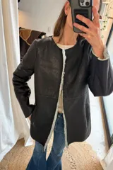 Campera Victoria de cuero genuino color negro, tipo mouton, con interior y bordes de corderito sintético en tono gris claro. Diseño de corte recto, sin cuello y con bolsillos laterales.