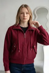 Campera de paño color marrón, con cuello alto, cierre frontal completo y cordones decorativos. Presenta bolsillos frontales con solapa y cierre a presión, y puños tejidos.