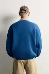 Sudadera azul con corte relaxed fit, cuello redondo y manga larga, confeccionada en tejido de algodón. Presenta un bordado frontal a contraste en color rojo con diseño ornamental y texto.