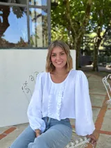 Blusa blanca de algodón con textura arrugada, cuello redondo, mangas largas abullonadas con puños elásticos y cierre frontal con botones y volantes.