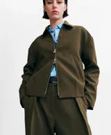 Chaqueta color verde militar, de corte recto con cierre frontal de cremallera, cuello camisero y bolsillos laterales.