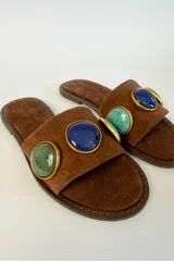 Sandalias planas de gamuza marrón con tira ancha decorada con dos apliques circulares de piedras en tonos verde y azul, enmarcados en metal dorado.