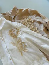 Camisa de manga larga en color blanco, confeccionada en tejido con textura. Presenta un diseño con bordado artesanal en relieve de color crema sobre uno de los hombros y parte del pecho, con cierre frontal de botones.