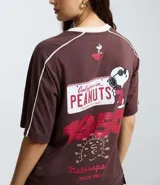 Remera de algodón color marrón con cuello redondo y mangas cortas. Presenta un estampado frontal con el personaje Snoopy en temática de carreras y detalles de líneas blancas contrastantes en los hombros y mangas.