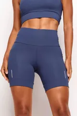 Short biker deportivo de tiro alto, confeccionado en tejido de poliamida con compresión moderada. Presenta bolsillos laterales funcionales, cintura anatómica con bolsillo interno 360° y detalles reflectivos en los muslos.