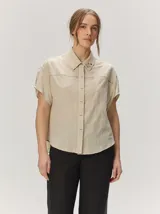 Camisa de manga corta color beige con sutil diseño a rayas verticales, cuello camisero con detalle de botones decorativos en la solapa y cierre frontal oculto.