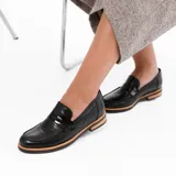 Mocasín de cuero vacuno negro con textura grabada tipo reptil. Presenta un diseño clásico con antifaz en el empeine, suela de goma en tono natural y taco bajo de madera.