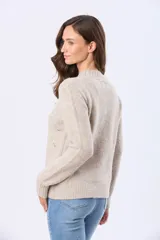 Buzo tejido de punto en color beige, con cuello redondo, mangas largas y puños acanalados. Presenta un delicado diseño bordado con motivos florales dispersos en el frente y una etiqueta de cuero sintético en el ruedo.