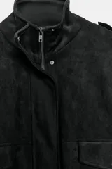 Cazadora bomber negra de gamuza, de cuello subido y manga larga con trabillas en los hombros. Cuenta con bolsillos de plastrón con solapa, cierre frontal con cremallera oculta y botones metálicos a presión, y elástico en el ruedo.