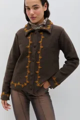 Chaqueta de lana merino hervida en color marrón, con diseño estructurado y cuello amplio. Presenta detalles de bordado artesanal en contraste color mostaza a lo largo del frente, cuello y puños, junto con botones forrados en la misma tela.