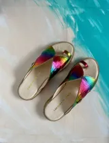 Sandalias de cuero vacuno modelo Anika de Carolina De Cunto, con diseño metalizado de efecto tornasolado en tonos arcoiris, tiras cruzadas y presilla para el dedo. Poseen suela plana y taco bajo de 2 cm.