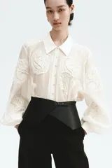 Blusa oversized de tejido transparente color crudo, con detalles bordados en relieve, cuello camisero, cierre frontal de botones y mangas largas abullonadas con puños ajustados.