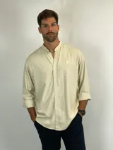 Camisa de manga larga en color beige, confeccionada en tejido de textura natural. Presenta cuello mao, cierre frontal con botones y un pequeño bordado distintivo en el pecho.
