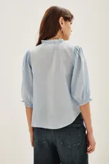 Camisa de algodón color celeste con cuello alto con volados, mangas tres cuartos abullonadas y cierre frontal con botones. Presenta un diseño con dos franjas verticales de bordado intrincado en color negro en el frente.