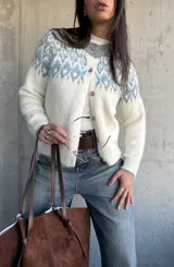 Cardigan de tejido de punto con diseño jacquard en la parte superior, cuello redondo y cierre frontal con botones. Presenta un patrón geométrico en tonos gris y azul sobre base color crema.
