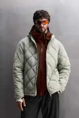 Campera puffer verde oliva acolchada con diseño de rombos, capucha y cierre frontal con cremallera. Incluye bolsillos laterales con cierre. Relleno de plumón y plumas.