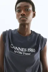 Remera de manga corta, cuello redondo y corte holgado, color gris oscuro o negro lavado, con estampado frontal en blanco que dice "CANNES 1980" y debajo "La Côte D'azur".