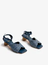 Sandalias de tacón medio en color azul, con capellada de rafia labrada en tonos azules y dorados, pulsera de cuero nobuck enlazada al tobillo con cierre de hebilla, y taco de madera natural lustrada de forma cilíndrica. Cuenta con puntera cuadrada.