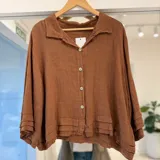 Camisa de lino color tostado, de corte holgado y corto, con cuello camisero, cierre frontal con botones y mangas largas con volados en los puños.