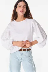 Blusa blanca de algodón, de corte recto y cuello redondo, con mangas largas abullonadas semitransparentes con textura calada de pequeñas flores y puños elásticos.