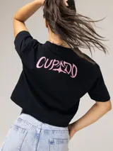 Remera crop negra Under Armour en colaboración con Tini, de manga corta y cuello redondo. Cuenta con un diseño gráfico frontal con el texto "FUTTTURA", fotos estilo polaroid y un listado de canciones en tonos rosados.