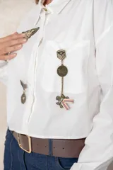 Camisa blanca de algodón, de corte corto y silueta holgada. Presenta cuello clásico, cierre frontal con botones y mangas largas con puños anchos. Está decorada con diversos parches y pines metálicos de estilo militar y náutico distribuidos en el frente y la manga derecha.