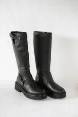 Bota de caña alta confeccionada en cuero negro, con suela gruesa tipo track antideslizante. Presenta un diseño con estribo integrado en el tobillo y una correa ajustable en la parte superior de la caña. Incluye cierre lateral para facilitar el calce.