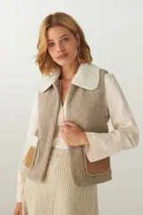 Chaleco corto de lana color beige, con cuello de piel sintética blanca y detalles de cuero marrón en los bolsillos.