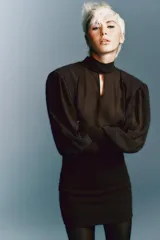 Vestido corto de color marrón con diseño combinado. Presenta un cuerpo superior fluido con cuello subido, lazada frontal ajustable y mangas largas abullonadas con hombreras y puños ajustados. La parte inferior es entallada con tejido elástico a tono, espalda con escote fluido y cierre lateral con cremallera oculta.