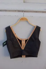 Top corto sin mangas, color negro, con escote pronunciado en V ribeteado en color beige. Presenta un cierre frontal con botones a presión plateados sobre una solapa central beige.