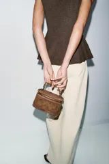 Bolso formato vanity de gamuza color marrón, con asa de mano superior y correa bandolera extraíble y ajustable. Presenta cierre de doble cremallera metálica dorada y detalles de pespuntes en el cuerpo.