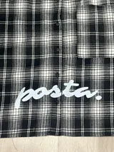 Camisa de manga larga con corte boxy fit, confeccionada en algodón a cuadros en tonos negro y gris. Presenta un bolsillo frontal en el pecho y el texto 'posta.' estampado en color blanco en la parte delantera.
