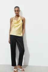 Top de cuello halter con escote fluido y espalda descubierta. Presenta un diseño satinado en color amarillo con estampado de lunares pequeños y un aplique metálico decorativo en el cuello.