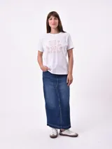 Remera de manga corta color beige claro con cuello redondo. Presenta un estampado gráfico central con un plato de pasta rodeado por un borde azul y blanco, sobre un fondo de franjas verticales rojas y blancas. El estampado incluye texto en cursiva en tonos turquesa y rojo.