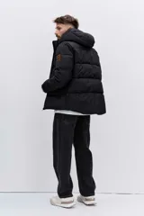 Cazadora acolchada negra con capucha, cierre frontal con cremallera y diseño de paneles horizontales. Presenta un parche decorativo en la manga y un ajuste cómodo.