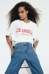 Remera blanca de algodón con estampado rojo de la frase "Los Angeles California U.S.A.", corte oversized, cuello redondo y mangas cortas.