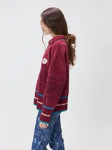 Pullover de textura tipo terciopelo con cuello alto y cierre frontal parcial. Presenta un diseño a rayas horizontales en tonos grises y negros, con detalles en rojo y blanco en el cuello, puños y ruedo. Incluye un aplique bordado con la letra M en el pecho y puños acanalados.