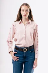 Camisa de algodón color rosado con cuello clásico y cierre frontal de botones. Presenta detalles de bordados florales en tonos rojizos a lo largo de las mangas y costuras con encaje decorativo en los hombros.