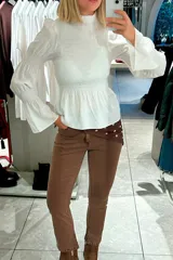 Blusa blanca de cuello alto con detalle de volado, cuerpo con fruncido elástico y mangas largas acampanadas con puños elásticos.