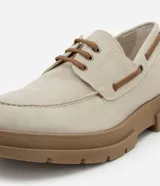 Zapato estilo dockside de color beige claro, confeccionado en gamuza sintética, con cordones marrones y suela track gruesa de goma color marrón. Presenta costuras visibles en la puntera y ojales metálicos para los cordones.