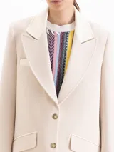 Saco sastrero estructurado confeccionado en sarga color beige, de largo por debajo de los muslos. Cuenta con cuello sastrero con solapa de pico ancha, broches dorados, hombreras, bolsillos con tapa y detalles en tela complemento en el cuello y bolsillos.