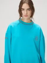 Buzo oversize color gris melange con corte crew, cuello redondo, mangas largas y detalles bordados en color verde en el cuello, puños y ruedo. Presenta el logo de la marca bordado en el pecho.