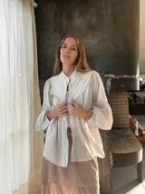 Camisa de algodón color beige con cuello mao, cierre frontal con botones y mangas abullonadas con frunces en los hombros. Presenta costuras verticales decorativas en el frente.